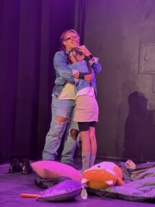 Two people hugging onstage with pillows on floor.